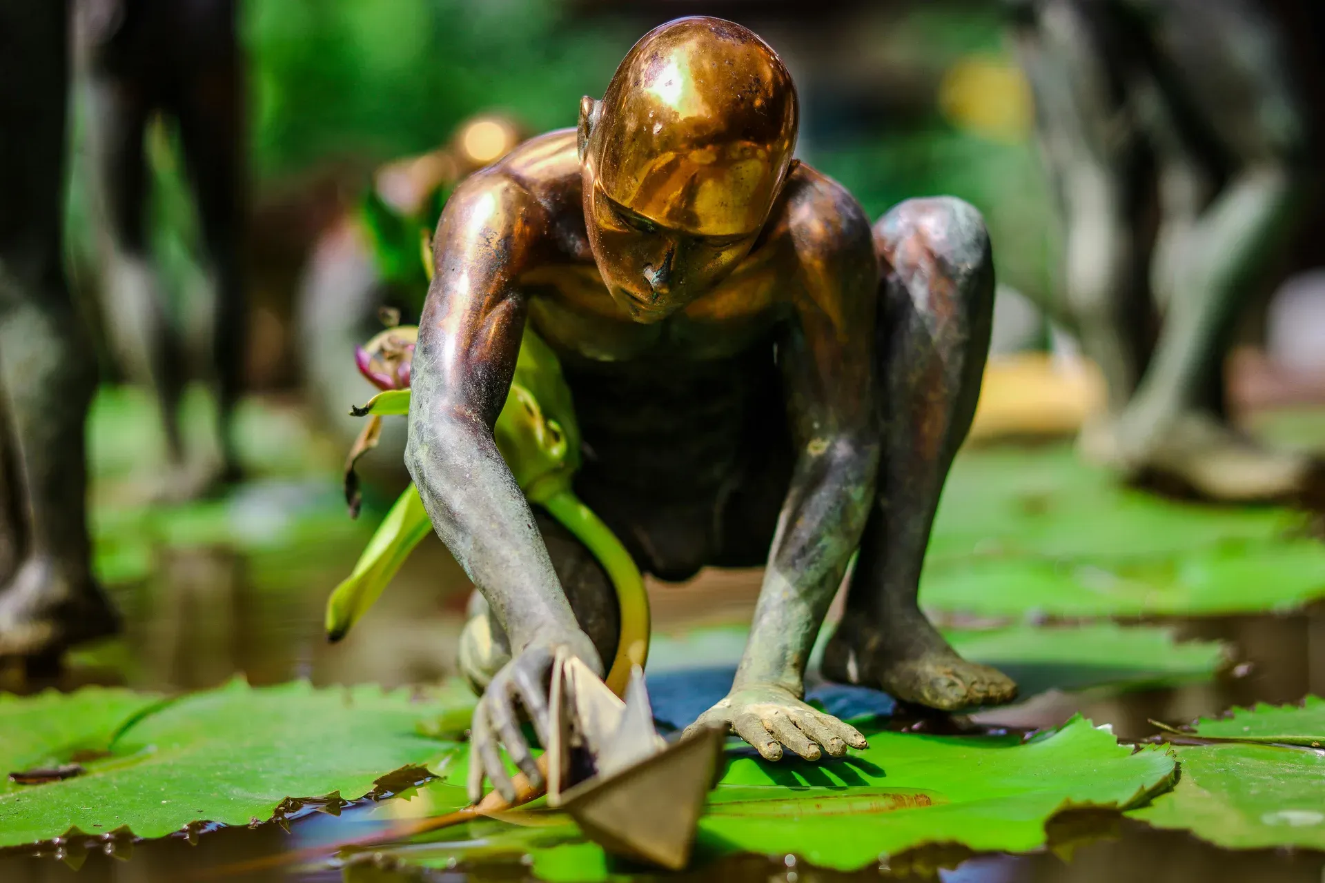 Escultura de bronce de figura agachada con cabeza dorada sobre nenúfares en un estanque