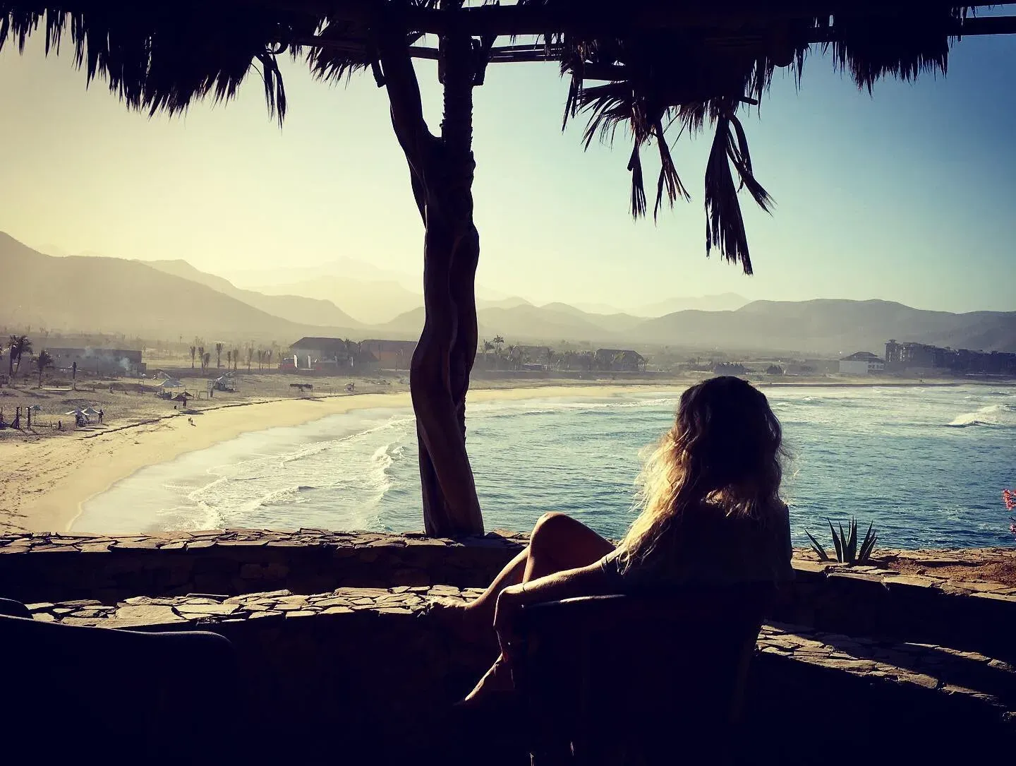 Mujer contemplando la playa desde una palapa con montañas al fondo