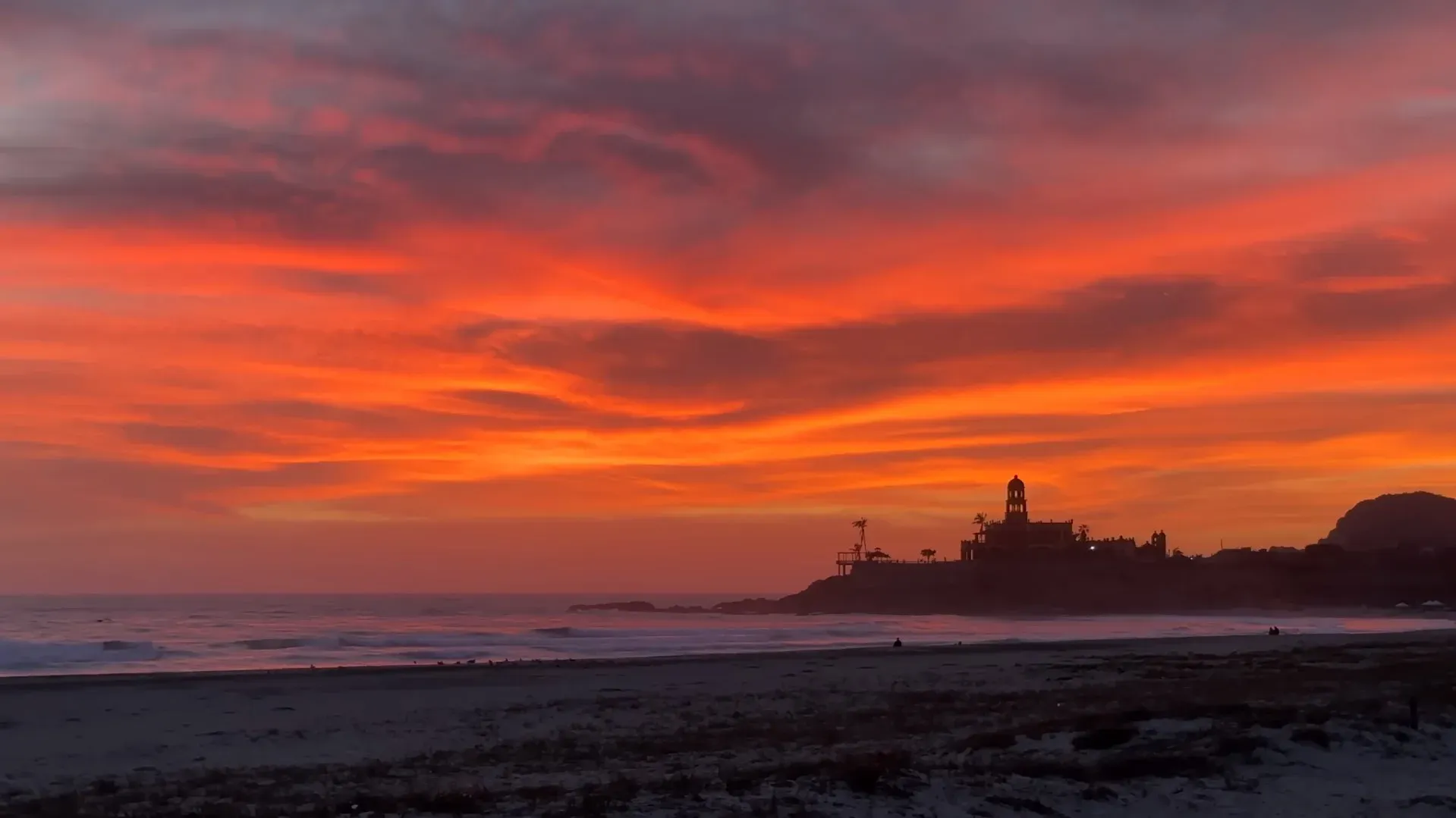 Atardecer rojo intenso en Playa Cerritos con silueta de edificio sobre el promontorio